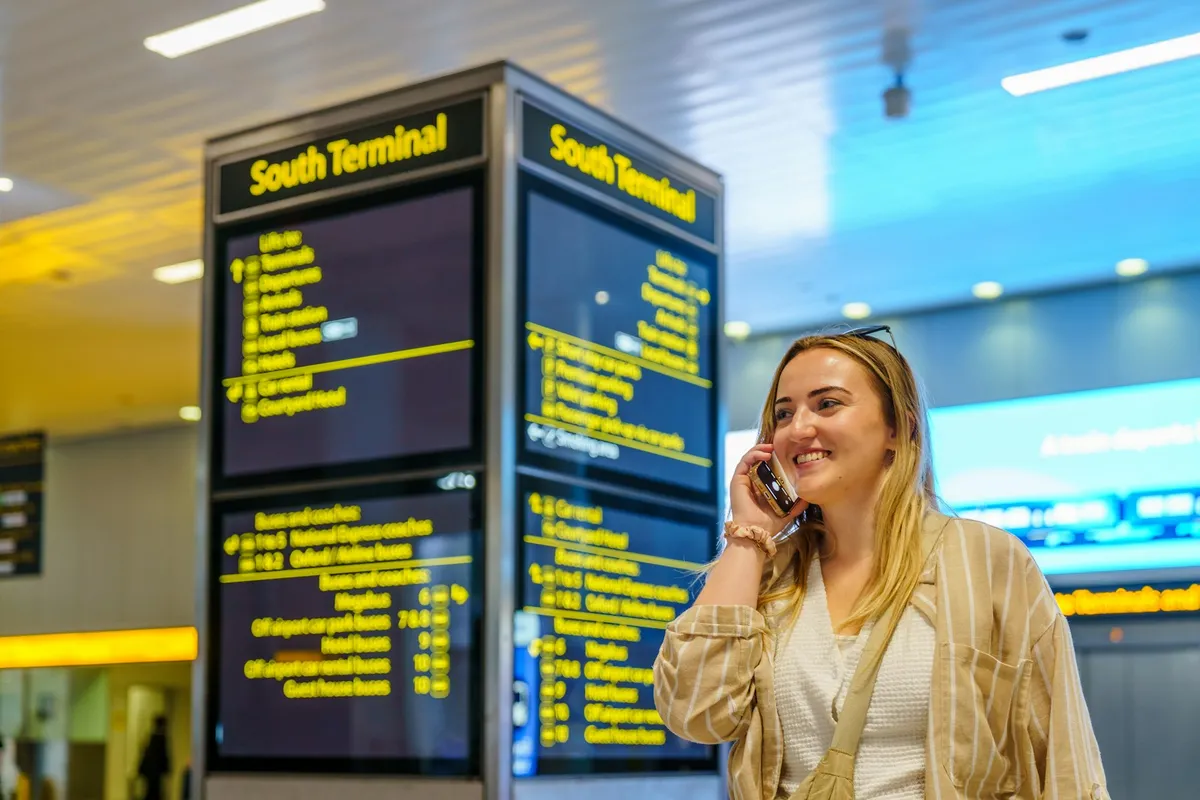 A smiling traveler on her phone standing in front of an airport departure information board.