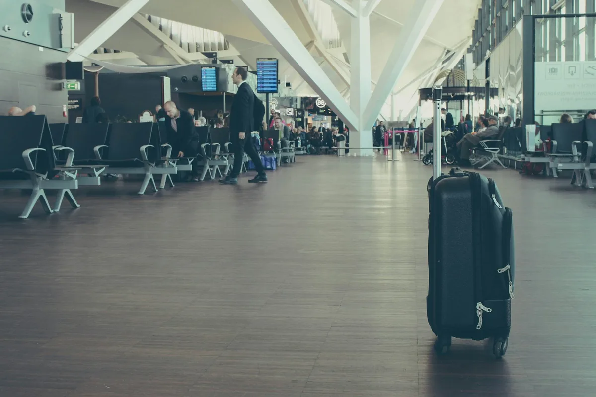 A black wheeled carry-on suitcase standing on the floor of a busy airport departure terminal