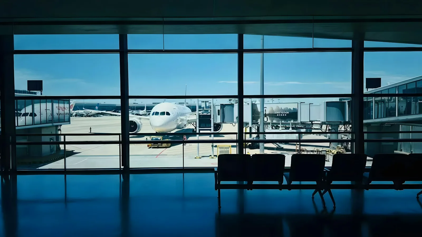 An airplane parked at a jet bridge seen through the floor-to-ceiling windows of an airport departure gate.