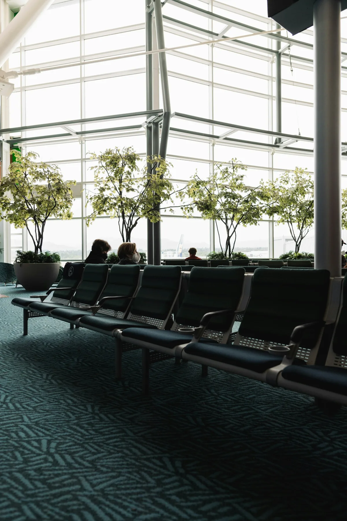 Passengers seated in an airport gate waiting area beside floor-to-ceiling terminal windows.