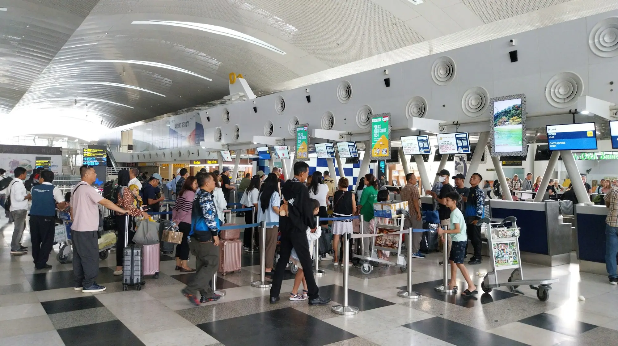 Travelers wait in line with luggage carts at an airport security and check-in area.